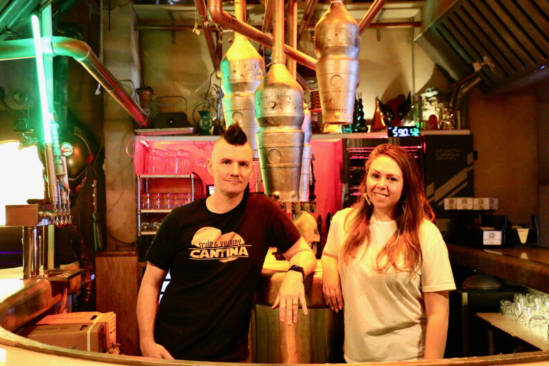 A light skinned man and woman stand behind a bar. The man has a mohawk haircut and is wearing a T-shirt that says Scum and Villainy Cantina. The woman has long red hair and is wearing a white T-shirt
