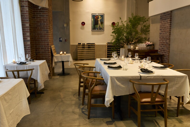 The interior of a restaurant shows a table set for eight people with a white tablecloth and modern wooden chairs on the right; around the room are other tables set for two. In the background is a modernist painting with red blue and green colors.