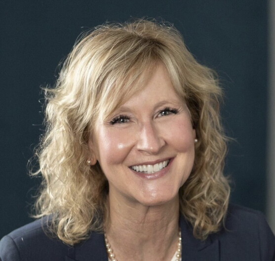 A woman with blonde, shoulder length hair, smiles while seated in front of a black background wearing a black blazer 