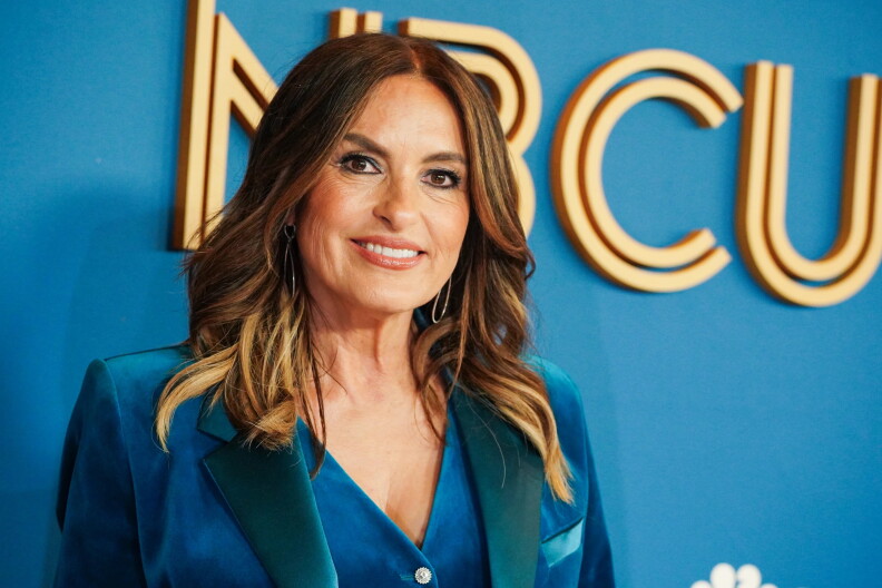 A woman wearing a bluse suit jacket and blue blouse stands in front of a blue background, smiling.