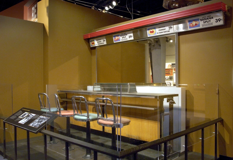 The lunch counter is an 8-foot section of the original lunch counter from Woolworth Department Store in Greensboro, North Carolina. There is a laminated black countertop with a stainless-steel trim along the front edge facing a line of four stools. A black, wooden, boxed footrest extends the whole length of the base of the counter.