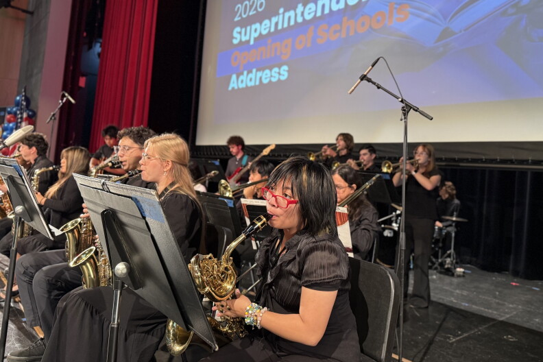 A teenage girl with dark brown hair and bangs wears red-framed glasses and a black, short-sleeved collared shirt and plays the saxophone. There are at least 10 other adolescents, also in black, playing various instruments, behind her.