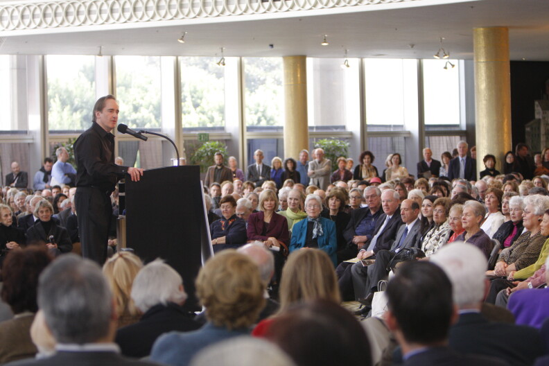 A male presenting person at a podium in front of a crowd of many people.