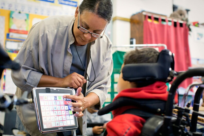 A woman in glasses helps a young boy in a wheelchair with navigating a computer screen.