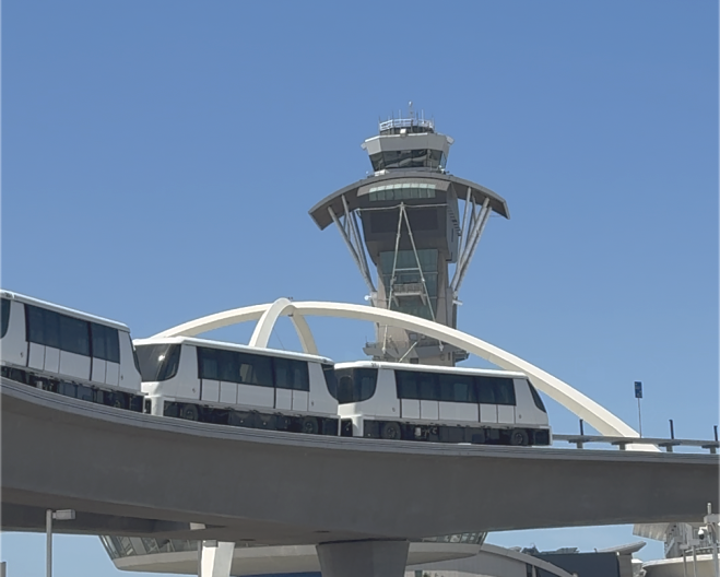 Three cars of a white train and black windows are visible on a gray track. There is a white arch behind the train. In the furthest background, there is a tower.