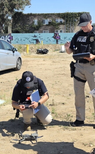 Two people are near a small grey drone that is on a dirt ground. One person is standing while holding a clipboard. Another is leaning over the drone and taking a photo on their phone. The dirt lot that they're standing on is vacant.