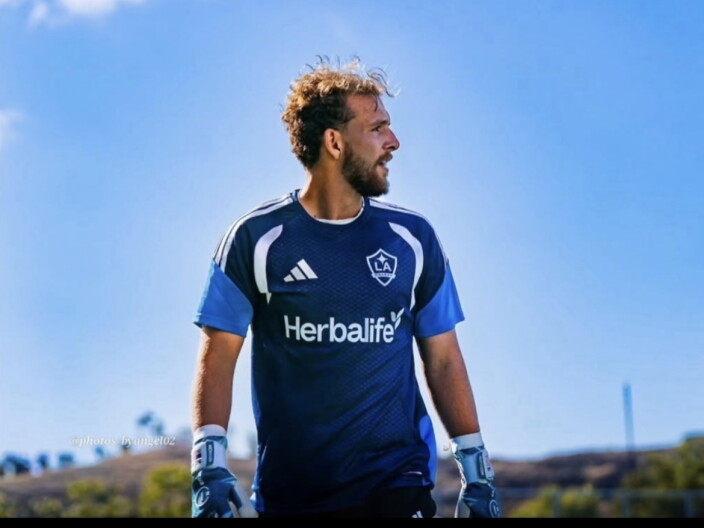 A light skinned male presenting person looks to the right. He wears a blue soccer team jersey and goal keeper gloves.