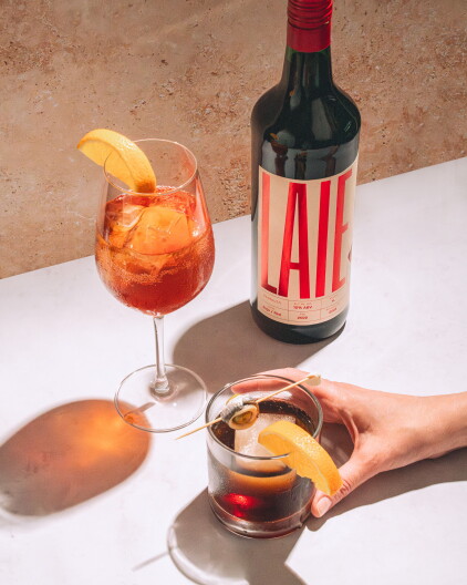 A bottle of LAIE vermouth beside two cocktails β a bubbly orange spritz and a dark vermouth over ice garnished with orange and an olive.