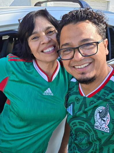 A man and woman both with medium-tone skin are wearing green Mexican soccer jerseys, and are smiling at the camera.