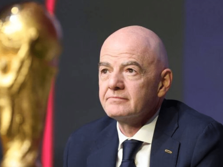 FIFA president Gianni Infantino with a neutral expression in suit and tie and in the foreground is a partially shown and blurred golden World Cup trophy