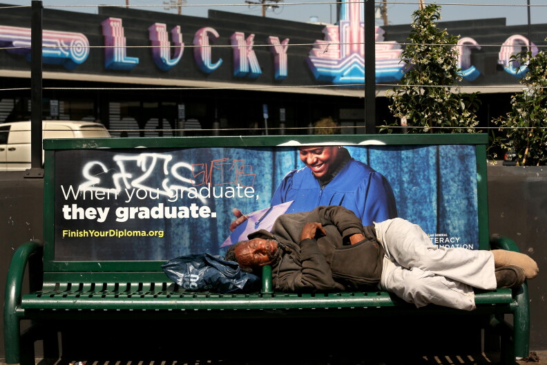 A man with dark skin tone sleeps on a green bus bench during daylight, using a backpack as a pillow. 