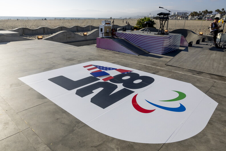 A view of an outdoor cement skate park near a beach, with a giant white logo that says "LA28" on it.