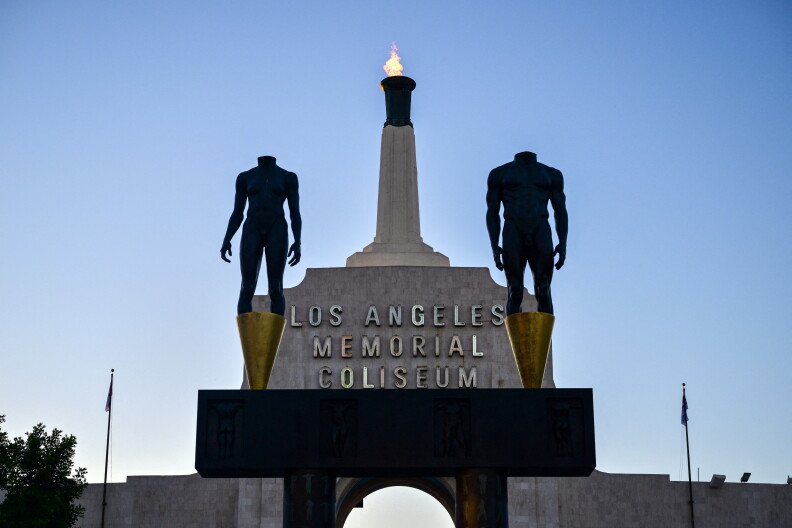 Two metal statues stand beside each other in front of a beige granite structure. Letters on the structure read "Los Angeles Memorial Coliseum" with a burning flag lit above it.