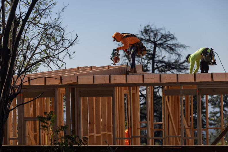 A wide look at two workers bent over on top of a wooden house frame using tools to work on it. One is wearing bright orange, while the other is in bright yellow.