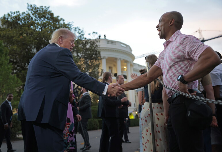 A heavyset man in a dark suit shakes hands with a dark-skinned man wearing a pink polo, in front of the White House rotunda. 