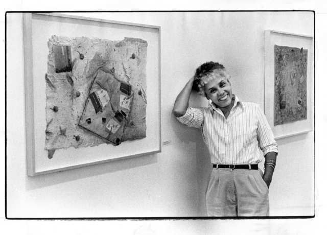 A Black woman in a shirt and slacks standing between two pieces of framed art pieces.