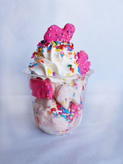 A sundae in a clear plastic cup topped with swirled whipped cream, rainbow sprinkles and pink and white frosted animal cookies, with pale pink ice cream visible below.