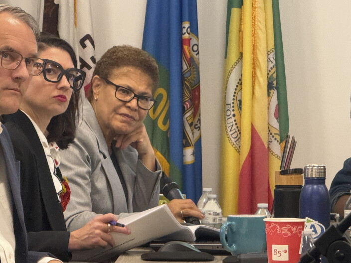 A woman with medium skin tone and short curly hair wearing black rimmed glasses and a gray blazer sits at a table with her head resting on her hand. Behind her are various flags.